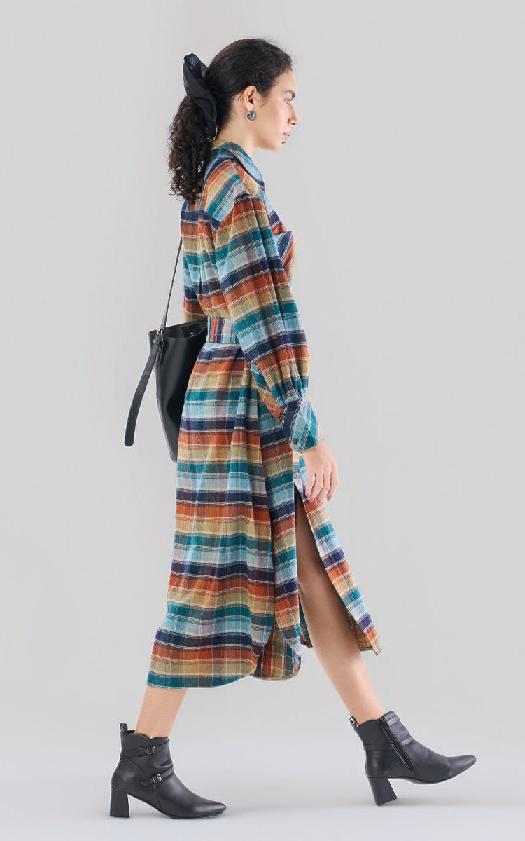 Woman wearing a long-sleeve multi-color plaid midi shirt dress in rust, navy, and teal with a waist-tie belt and black leather ankle boots.